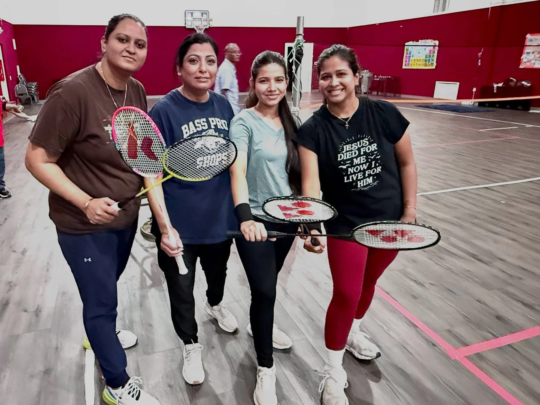 Four women in casual sportswear stand together on an indoor badminton court, smiling and holding their rackets towards the camera.