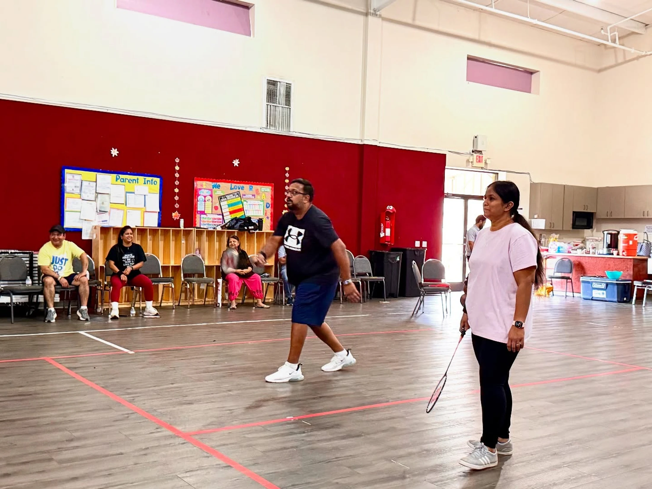 A man and a woman play badminton on an indoor court with a wood floor as other people watch from chairs in the background.