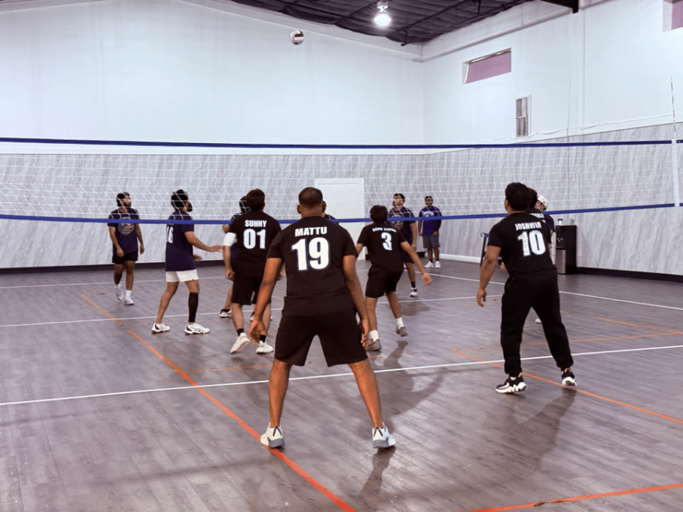 A group of men playing volleyball on an indoor court. The ball is in the air over the net, and players are in position, ready for the next move.