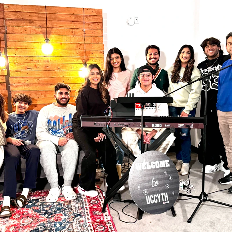 A group of ten smiling young people pose for a photo. A man sits at a keyboard in the center, and a sign in front reads 'Welcome to UCCYTH'.