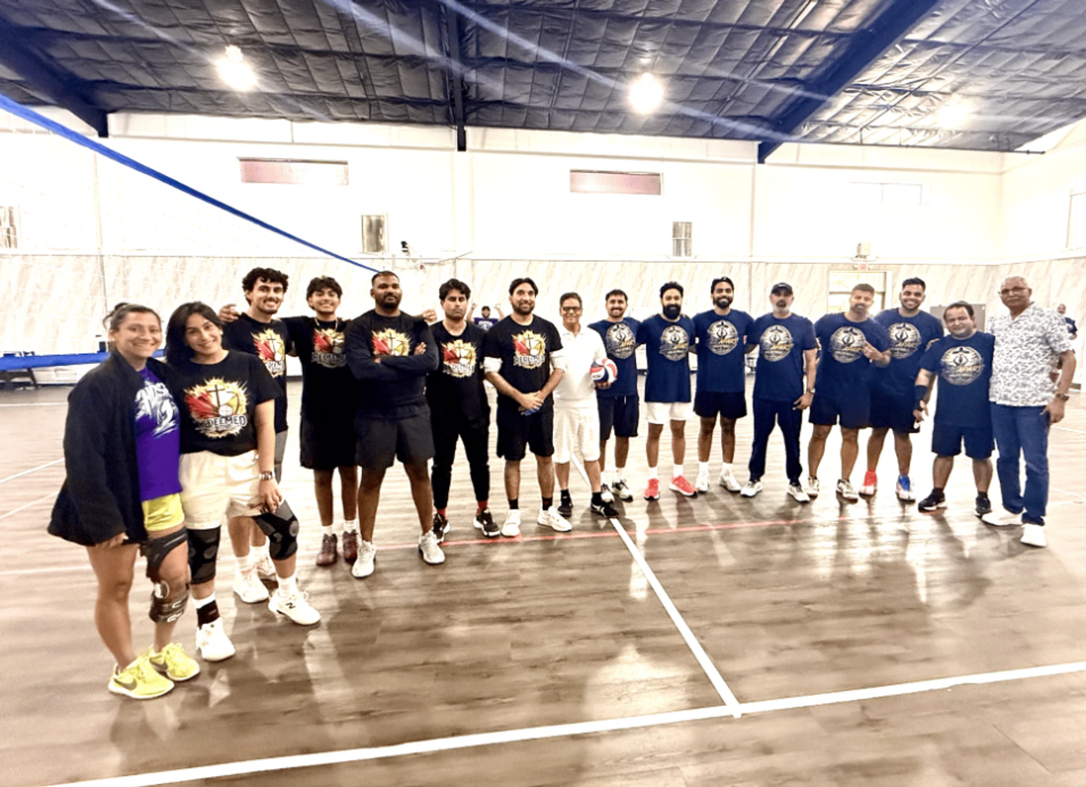 A large group photo of two volleyball teams, one in black shirts and one in blue, standing together and smiling on an indoor gymnasium court.