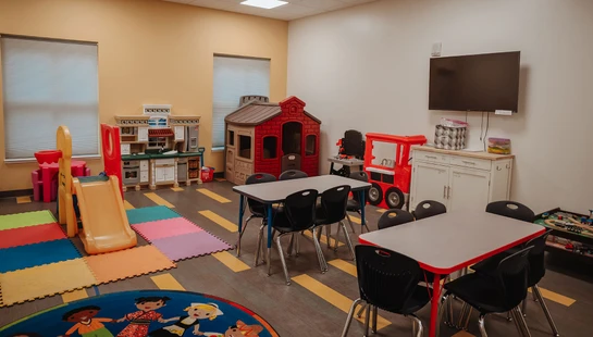A brightly lit playroom for children with small tables, chairs, a slide, a play kitchen, a playhouse, and other colorful toys on a patterned floor.
