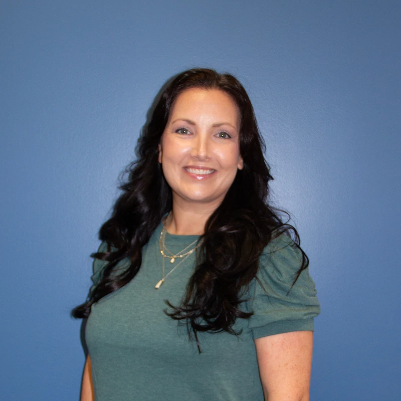 A smiling woman with long, dark, wavy hair poses for a headshot in front of a solid blue background. She is wearing a green shirt and a layered necklace.