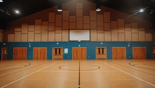 A wide shot of an empty gymnasium with a polished basketball court, facing a blue wall with several wooden doors and a large projector screen.