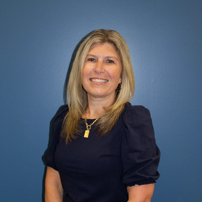 A smiling woman with blonde, shoulder-length hair poses for a headshot against a solid blue background. She is wearing a navy blue top and a gold necklace.