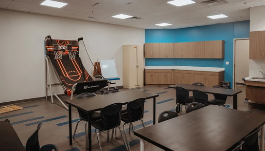 A brightly lit classroom or activity room with dark tables and chairs. In the corner is an arcade basketball game, and a blue wall has wooden cabinets.