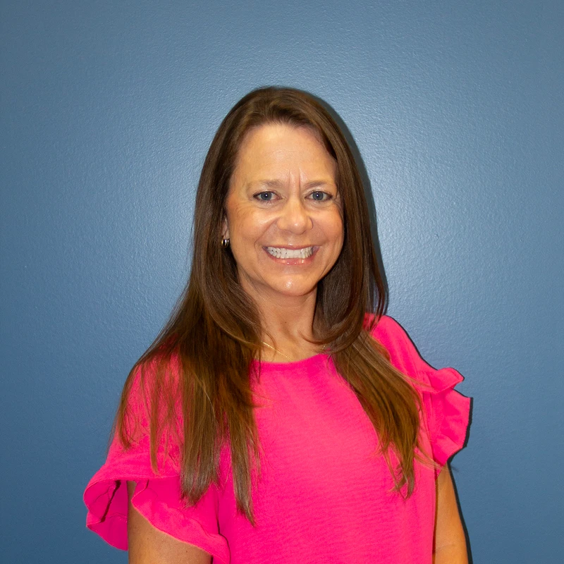 A head and shoulders portrait of a smiling woman with long brown hair and blue eyes, wearing a vibrant pink top against a solid blue background.
