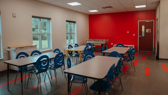 An empty classroom with tables, blue swirl-backed chairs, a foosball table, and a bright red accent wall. Red markers are on the floor.