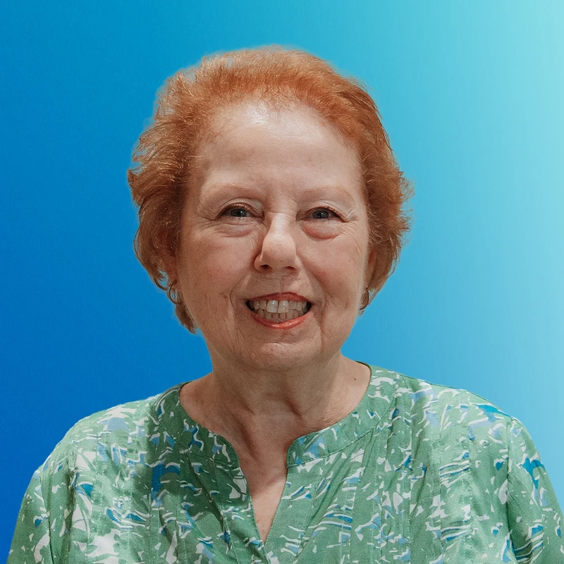 A headshot of a smiling older woman with short red hair. She is wearing a green patterned blouse and is set against a bright blue gradient background.