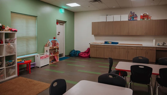 An empty children's classroom with light green walls, featuring tables and chairs, shelves full of toys, a dollhouse, and a long counter with cabinets.