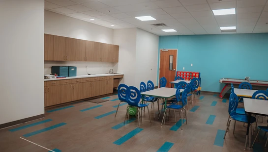 A modern children's activity room featuring small tables with unique blue chairs, a teal accent wall, wooden cabinetry, and large floor games.