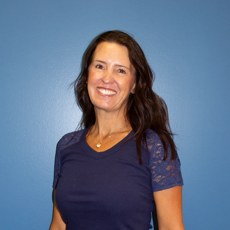 A head-and-shoulders portrait of a smiling woman with long brown hair, wearing a blue shirt with lace sleeves, against a solid blue background.