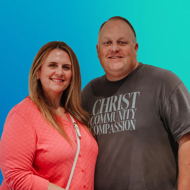 A smiling man and woman stand side-by-side against a blue and green gradient background. The man wears a t-shirt that says 'Christ Community Compassion'.