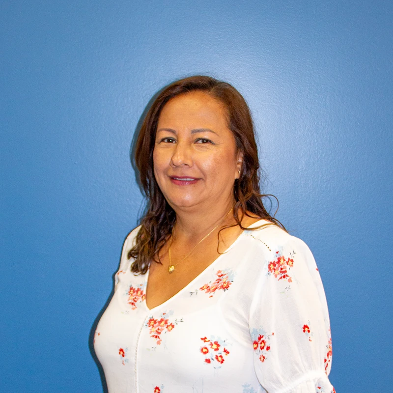 A head and shoulders portrait of a smiling woman with brown hair, wearing a white floral blouse in front of a solid blue wall.