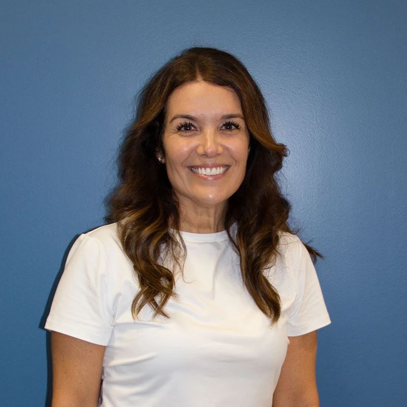 A headshot of a smiling woman with long brown hair, wearing a white t-shirt and standing in front of a solid blue wall.