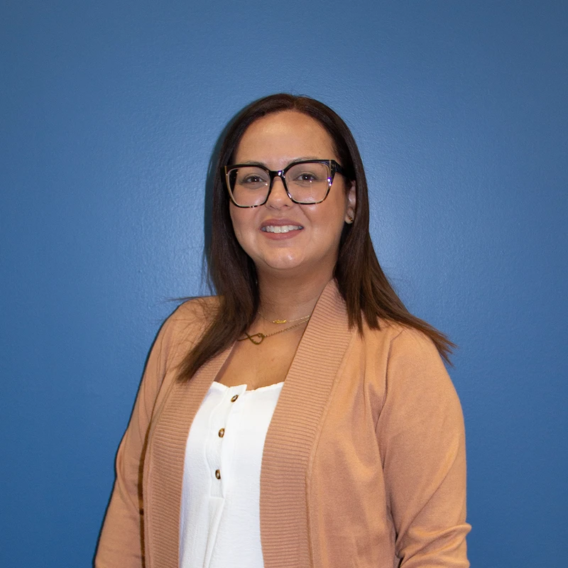 A woman with long brown hair and black glasses smiles at the camera. She is wearing a white shirt and a peach cardigan, standing before a blue background.