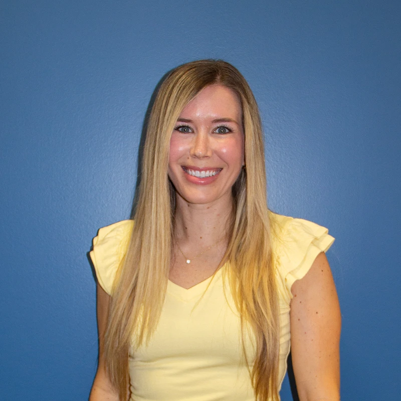 A head-and-shoulders portrait of a woman with long blonde hair, smiling at the camera. She wears a yellow shirt and stands before a solid blue background.