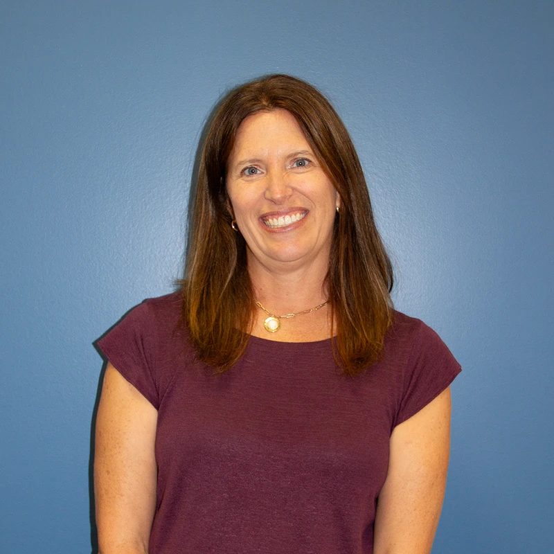 A smiling woman with long brown hair and blue eyes wearing a maroon shirt and a gold necklace, posing for a headshot against a blue background.