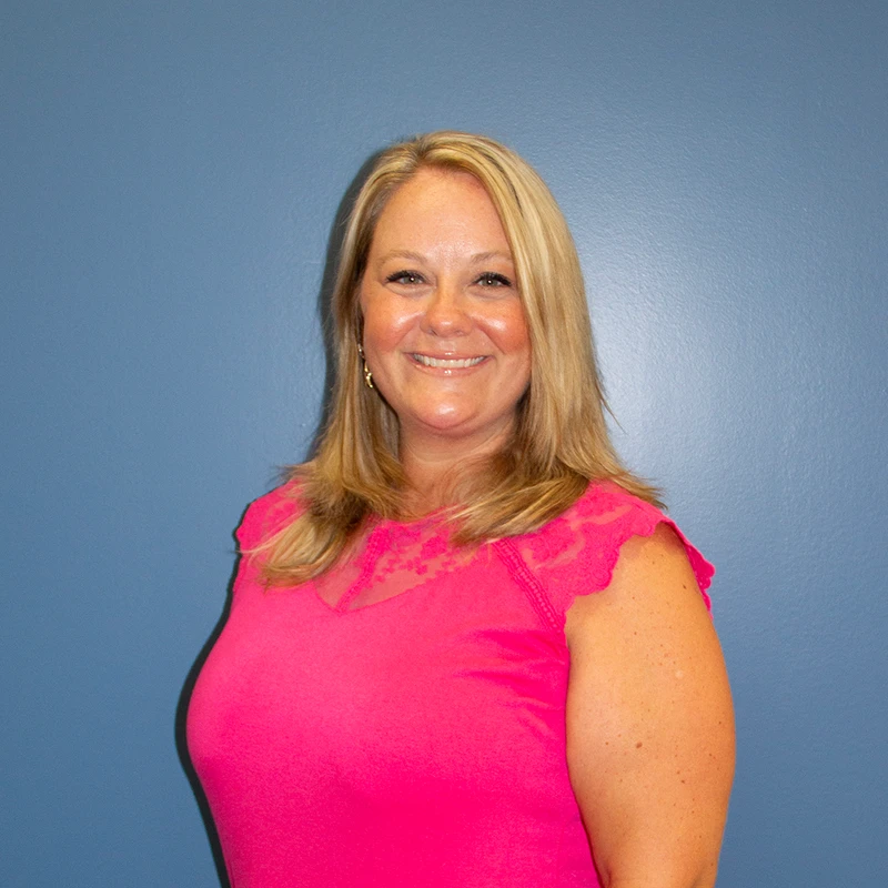 A professional headshot of a smiling woman with blonde hair. She is wearing a bright pink top and standing in front of a solid blue wall.