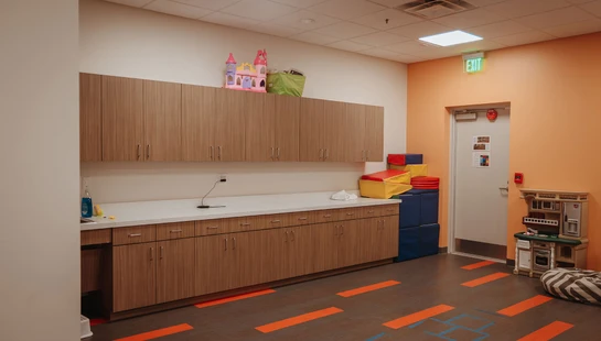 A brightly lit children's playroom with an orange wall, wood cabinets, a toy kitchen, soft play blocks, and orange markings on the floor.