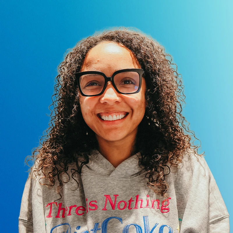 A close-up portrait of a smiling woman with long curly hair and black glasses, wearing a grey hoodie against a solid bright blue background.