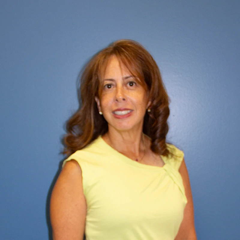 A head and shoulders portrait of a woman with shoulder-length brown hair, smiling at the camera. She wears a yellow sleeveless top against a blue background.