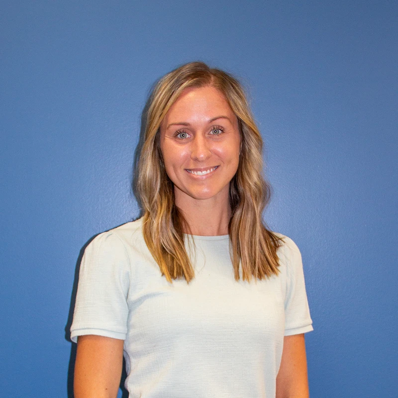 A professional headshot of a woman with blonde hair smiling at the camera. She is wearing a light green shirt and stands against a solid blue background.