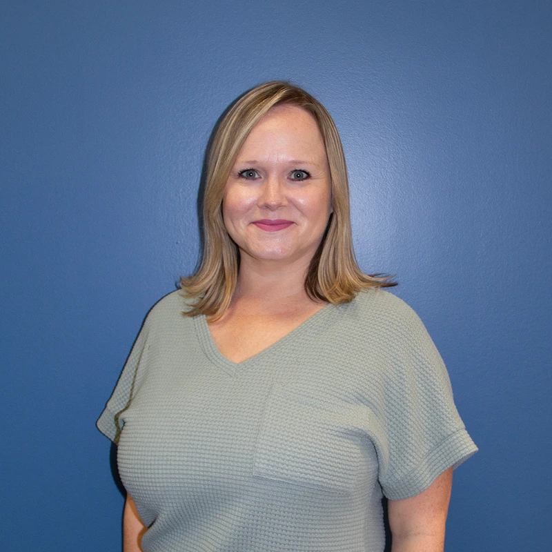 A smiling woman with shoulder-length blonde hair poses for a headshot in a sage green top against a solid blue background.