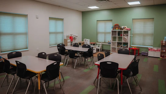 An empty children's classroom or daycare with small tables, chairs, and various play areas with toys organized along the walls.