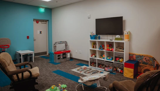 A brightly lit nursery room with a teal wall, gray carpet, rocking chairs, and shelves filled with colorful toys for children. A TV is mounted above the shelf.
