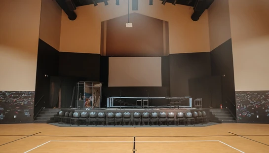 A wide shot of an empty multipurpose room with a basketball court floor, facing a raised stage set with chairs, microphones, and musical instruments.