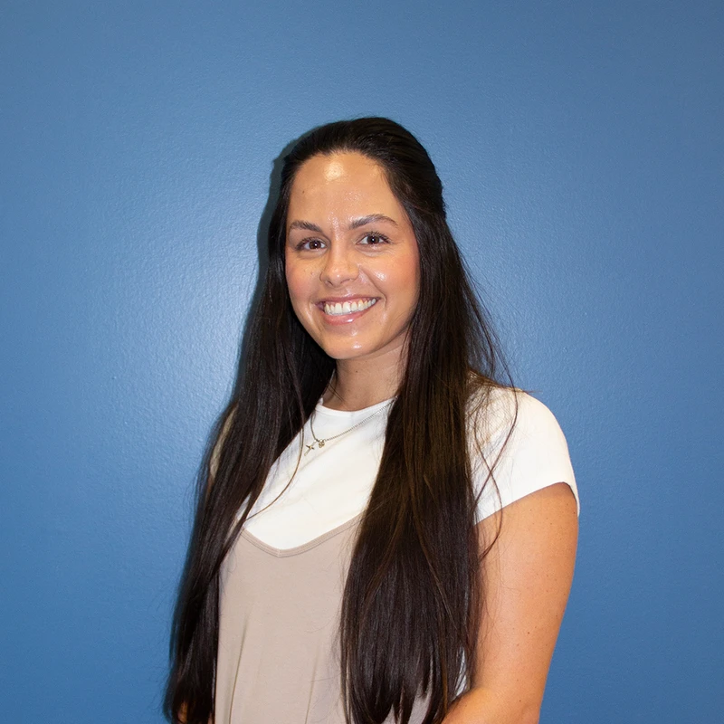 A smiling woman with long, dark brown hair stands in front of a solid blue wall. She is wearing a white shirt under a beige top.