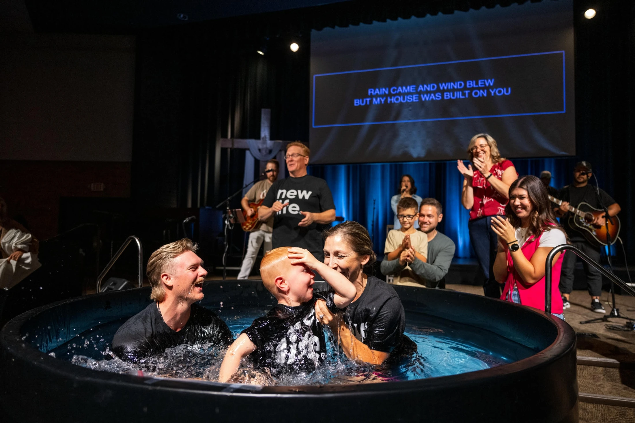 A young boy smiles as he is baptized by a man and a woman in a tub on a church stage. The congregation and a band look on and clap in celebration.