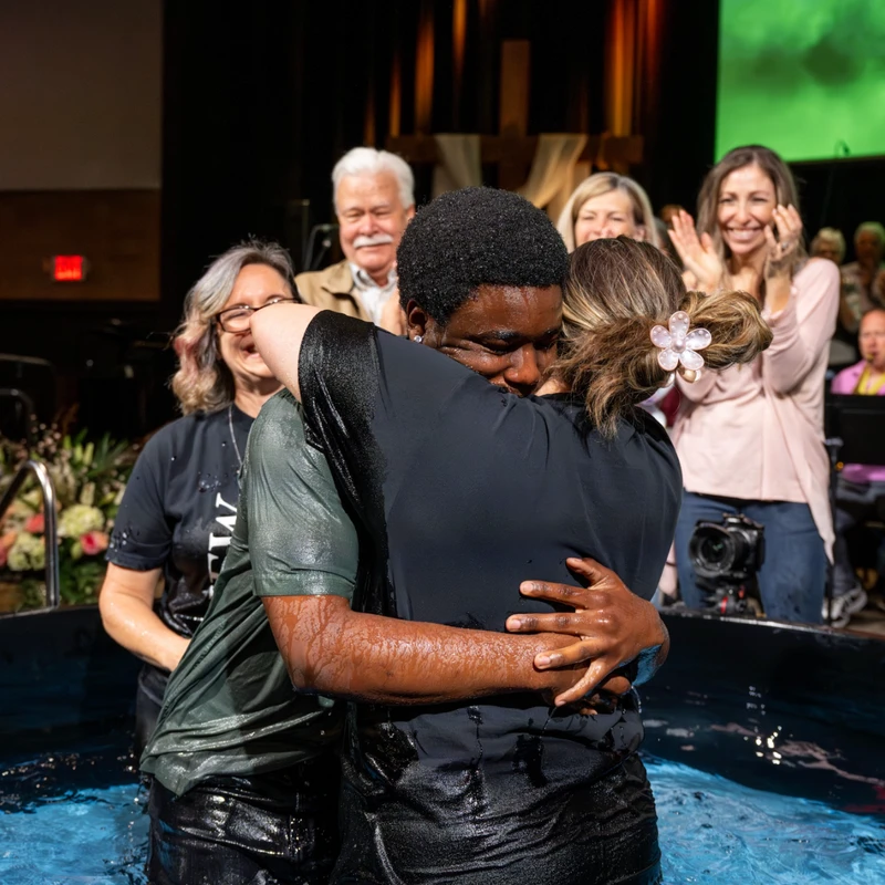 A man and a woman, soaking wet, share an emotional hug in a baptismal font as onlookers smile and clap for them in a church.