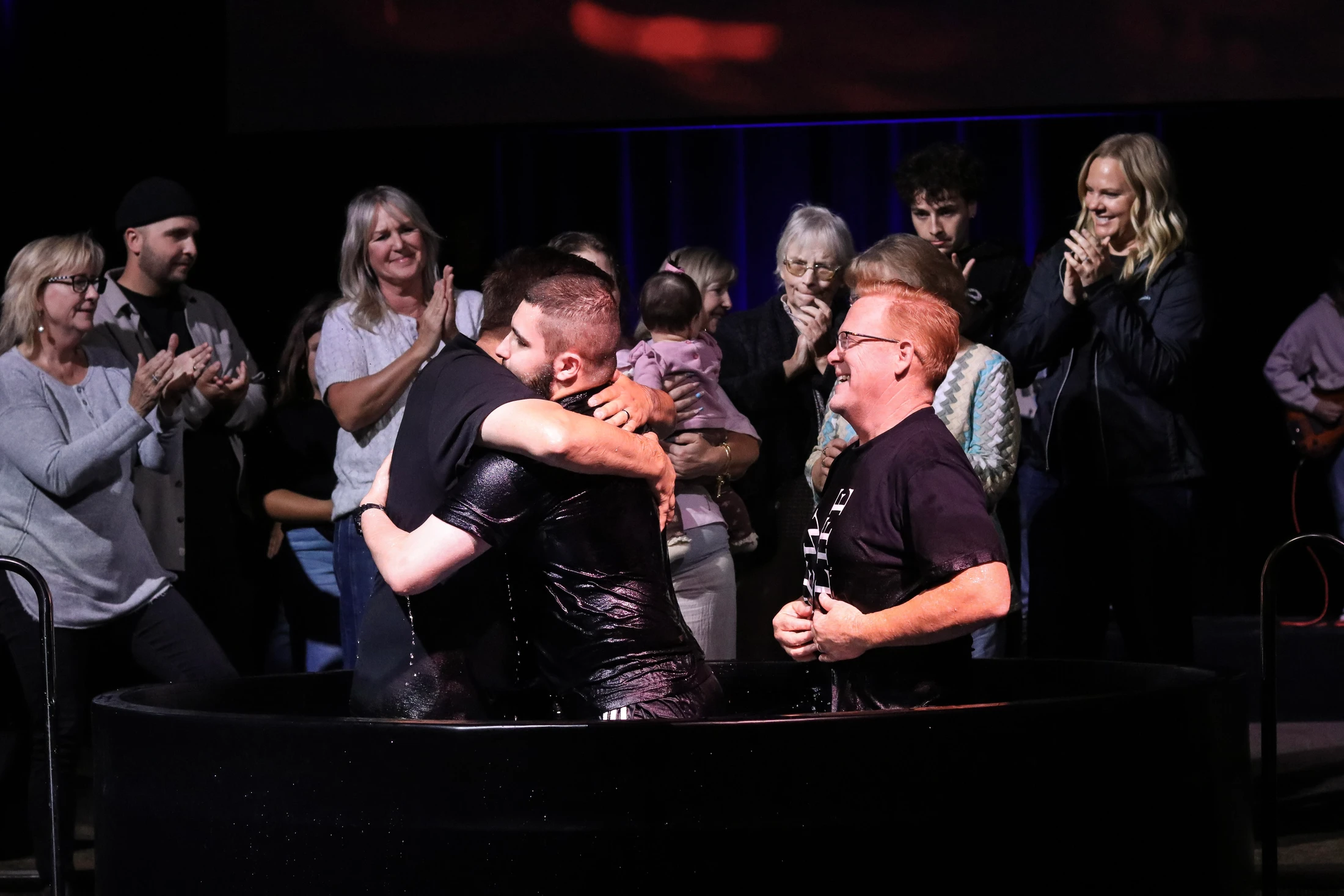 Two men embrace in a baptismal font after a baptism ceremony, while a pastor smiles and a crowd of onlookers applauds in celebration on stage.
