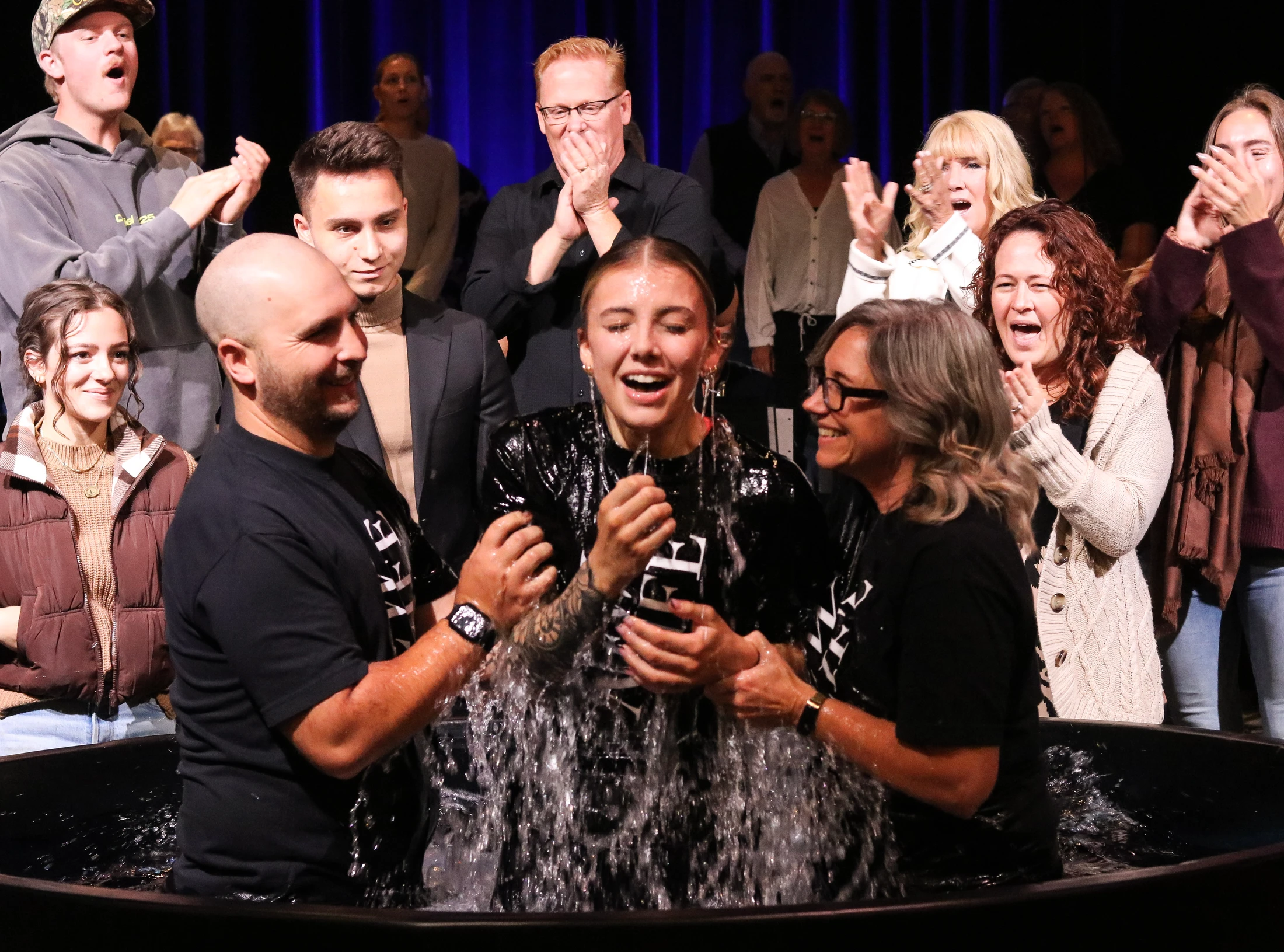 A young woman with a joyous expression emerges from the water of a baptismal font, supported by a man and woman. A crowd watches and applauds from the stage.