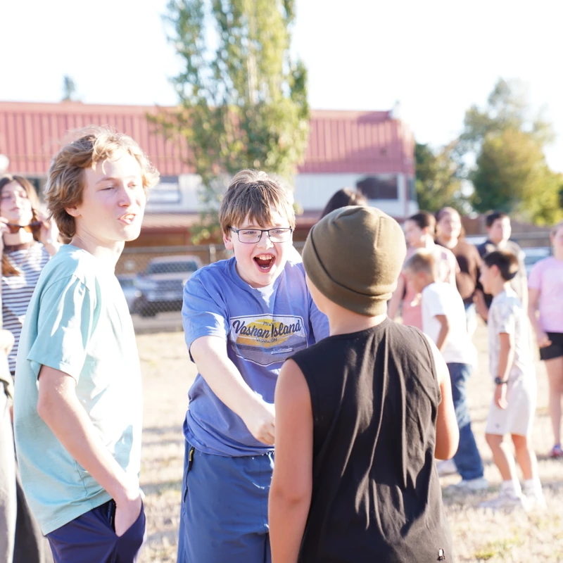 A group of young people stand in a grassy field. In the foreground, a boy with glasses and a blue shirt laughs with an open mouth while talking to two friends.