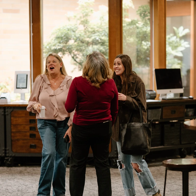 Three women stand together in a well-lit room, engaged in a lively conversation. One woman laughs, another smiles, creating a joyful, candid moment.