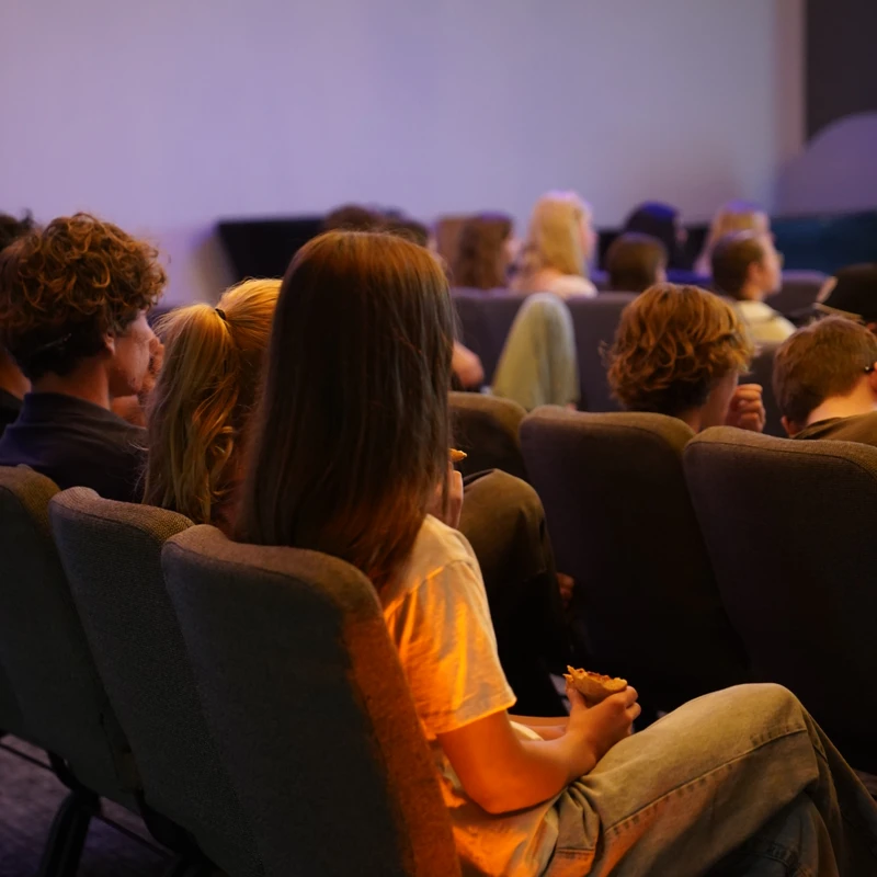 A group of young people seen from behind, sitting in rows of chairs in a dark room and looking towards a large, illuminated screen at the front.