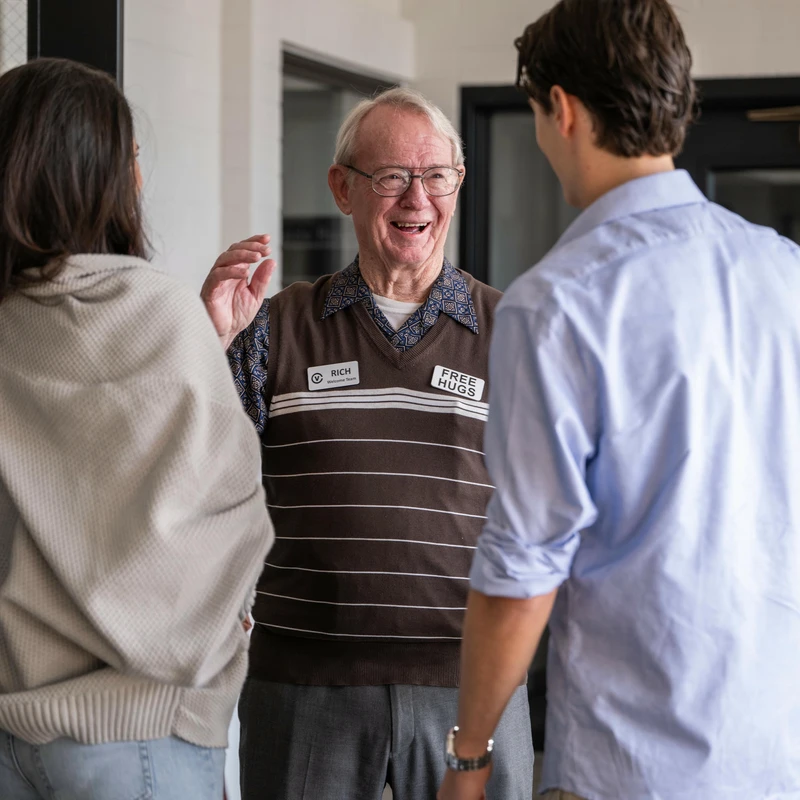 A friendly elderly greeter named Rich, wearing a 'Free Hugs' tag, smiles and talks with a young man and woman in a building lobby.
