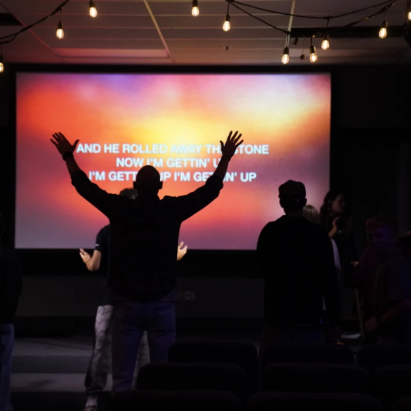 Silhouette of a man with arms raised in worship, facing a large screen with song lyrics during a dimly lit church service with a band on stage.
