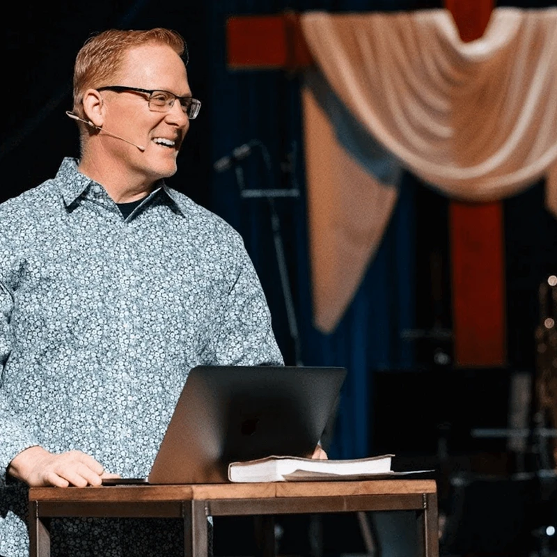 A man with red hair and glasses smiles while standing behind a lectern on stage. He wears a headset microphone and a patterned shirt.