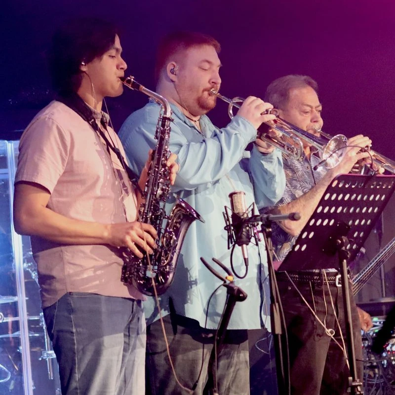Three male musicians stand side-by-side on a dimly lit stage, playing a saxophone and two trumpets while reading from a music stand.