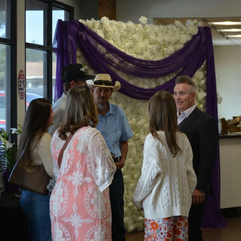A group of people stand in a circle talking at an event. Behind them is a decorative wall covered in white flowers and draped with purple fabric.
