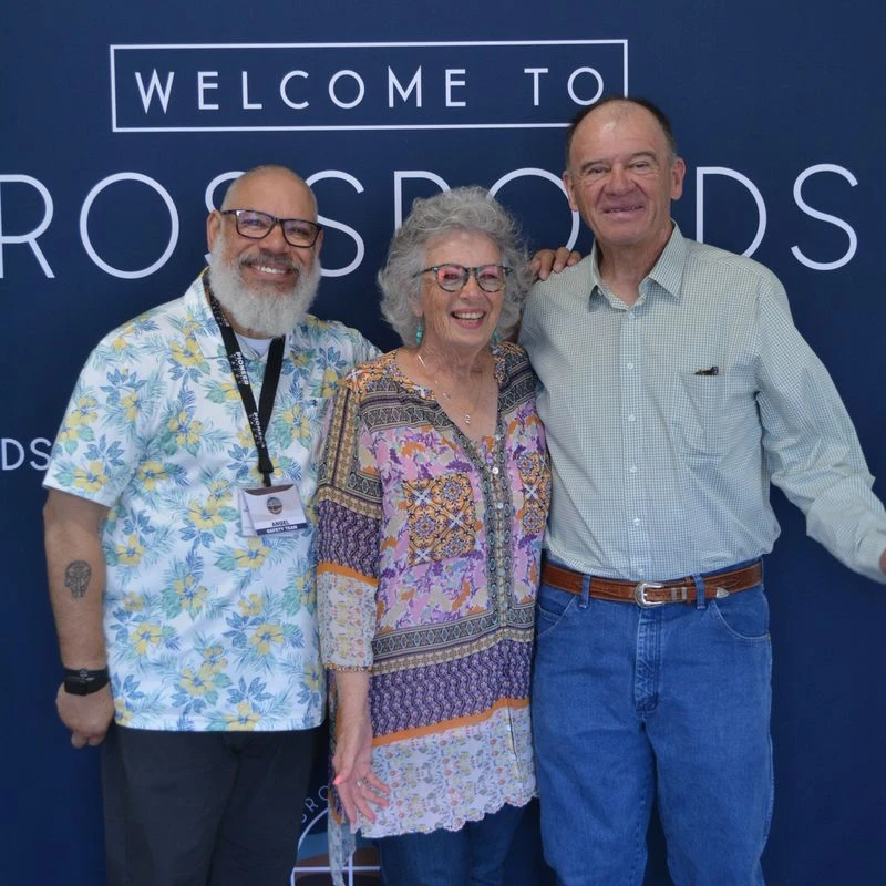 Two men and a woman stand arm-in-arm, smiling for a photo in front of a large blue banner that reads, 'WELCOME TO CROSSROADS.'