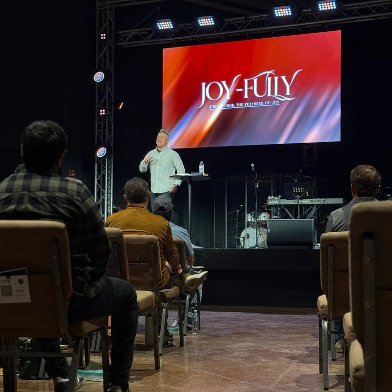 A pastor preaches on stage to a seated audience. A large screen behind him reads 'JOY-FULLY, Discovering the Fullness of Joy'.