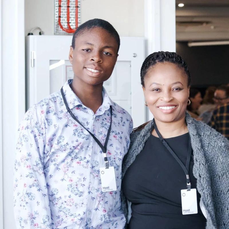 A young man and a woman stand side-by-side, smiling for the camera. They both wear lanyards with badges that identify them as 'Host Guest Services'.
