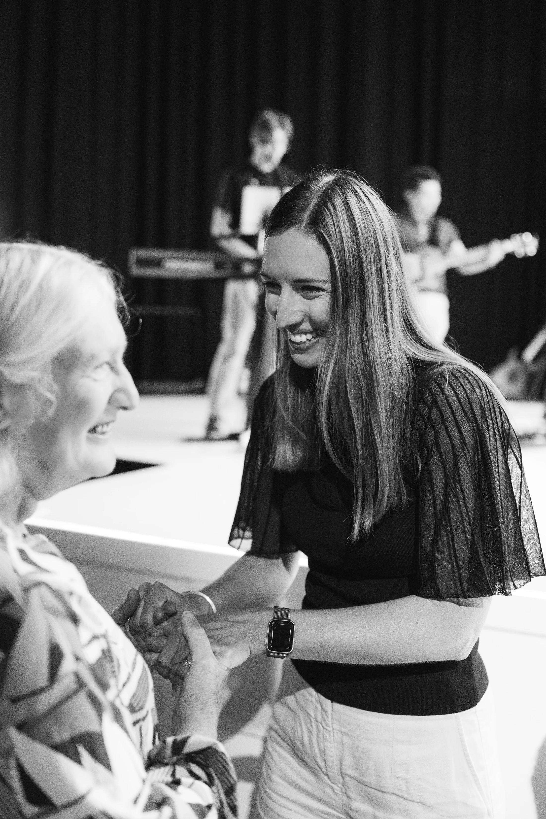A black and white photo of a smiling young woman holding the hands of an older woman. They are looking at each other warmly, with musicians in the background.