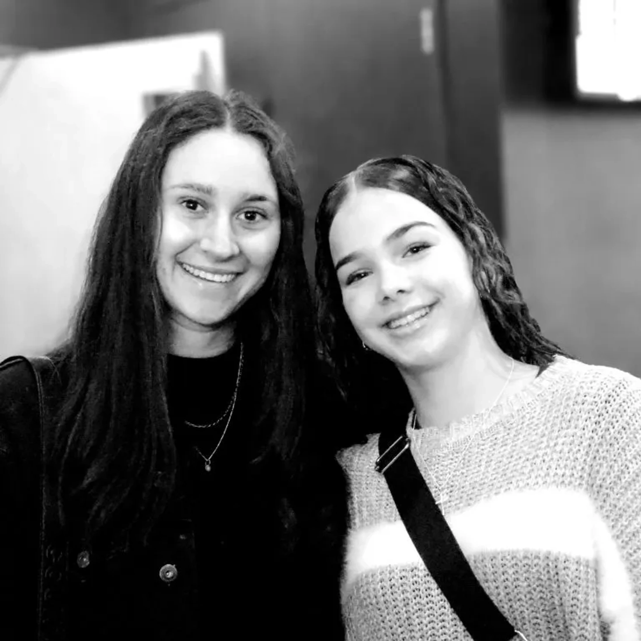 A black and white photo of two young women with long dark hair smiling for the camera. One wears a dark top and the other wears a striped sweater.
