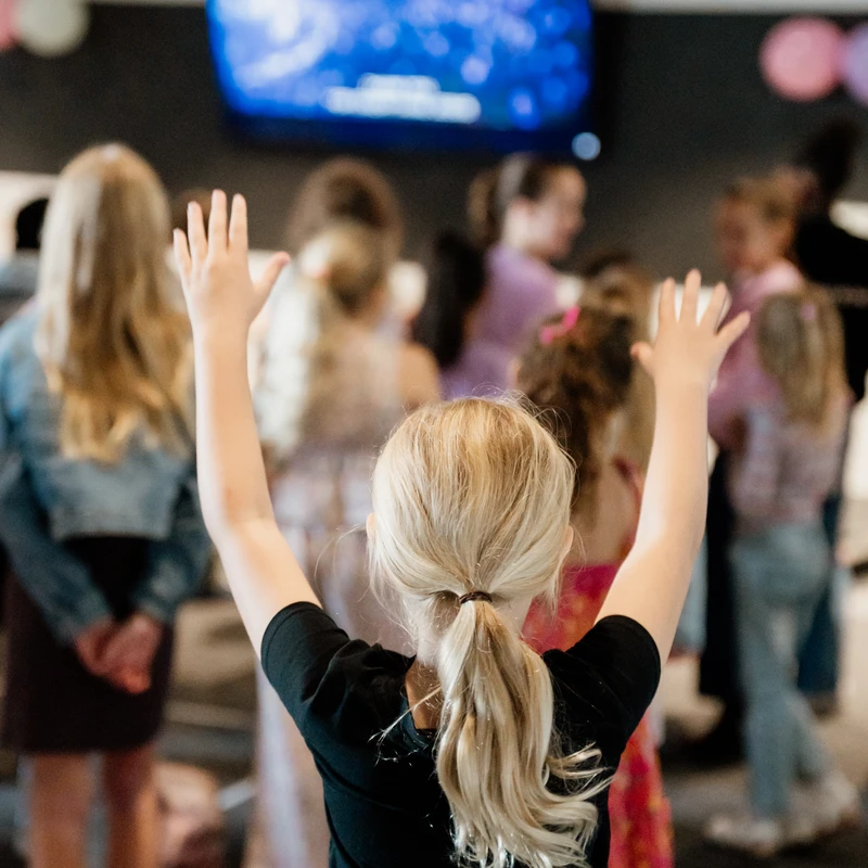 A young girl with blonde hair in a ponytail, seen from behind, raises her hands in the air. She is in a room full of other children watching a screen.